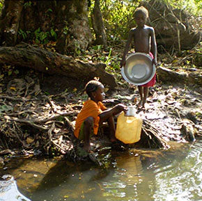 Two children collecting water from a stream using a metal bowl and a plastic jug